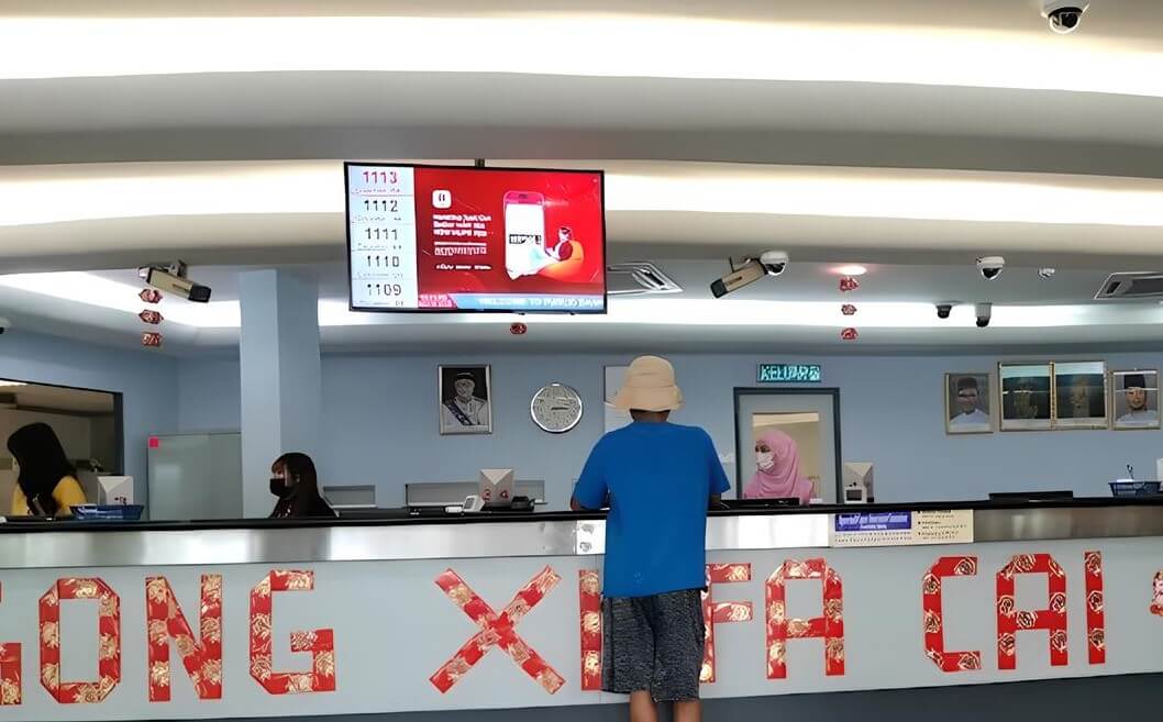 Seorang pelanggan yang memakai topi matahari kuning air dan baju biru berdiri di kaunter bank Public Bank Parit Raja, membelakangi kamera. Sepanduk merah yang tertera perkataan "GONG XI FA CAI" dalam warna emas tergantung di kaunter. Di belakang kaunter terdapat beberapa juruwang bank, salah seorang daripadanya memakai tudung merah jambu