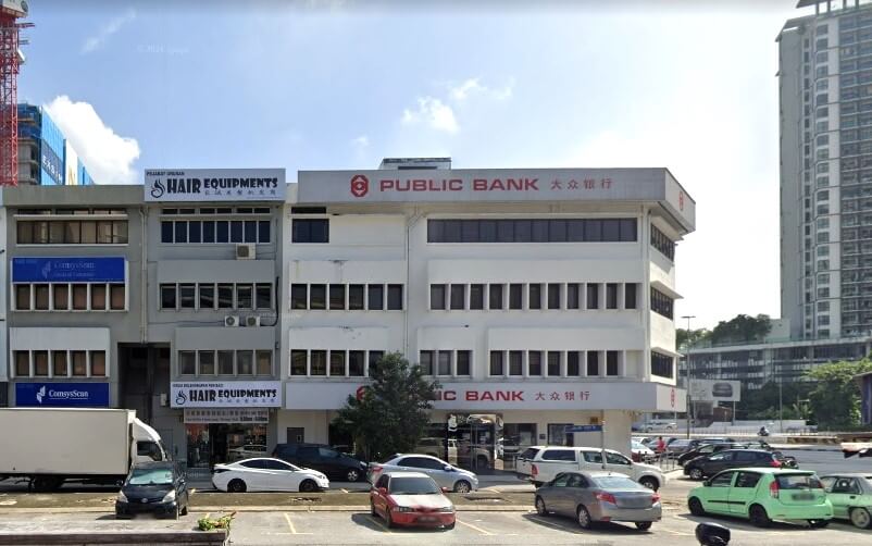 Pemandangan jalan waktu siang menunjukkan deretan bangunan komersial bertingkat rendah. Bangunan pusat ialah struktur putih empat tingkat dengan logo dan nama Public Bank dalam kedua-dua aksara Inggeris dan Cina. Di kiri terdapat dua bangunan kelabu yang bersambung. Di latar belakang paling kanan, bangunan pencakar langit moden yang tinggi, dalam pembinaan atau baru siap, menguasai latar langit