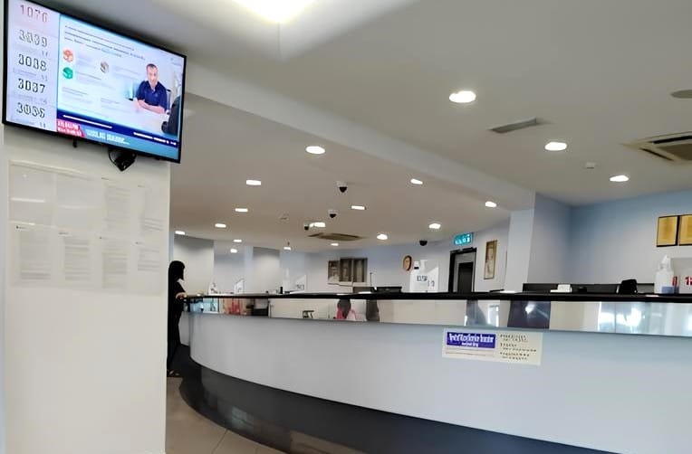 Bahagian dalam cawangan Public Bank di Jalan Kuchai Lama. Di latar depan, terdapat kaunter perkhidmatan melengkung yang besar dengan reka bentuk kelabu putih dan gelap. Papan tanda kecil berwarna biru dan putih, mungkin mengandungi maklumat perkhidmatan pelanggan, dilampirkan di hadapan kaunter. Di sebelah kiri, dipasang pada tiang atau dinding, ialah skrin digital yang memaparkan sistem baris gilir