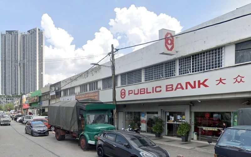 Pemandangan jalan yang menunjukkan muka bangunan cawangan Public Bank di Segambut, Malaysia. Bangunan dua tingkat itu mempunyai bahagian luar berwarna putih dengan perkataan 'PUBLIC BANK' dalam huruf merah besar, bersama-sama aksara Cina. Kompleks perumahan pencakar langit moden yang tinggi kelihatan di latar belakang di sebelah kiri
