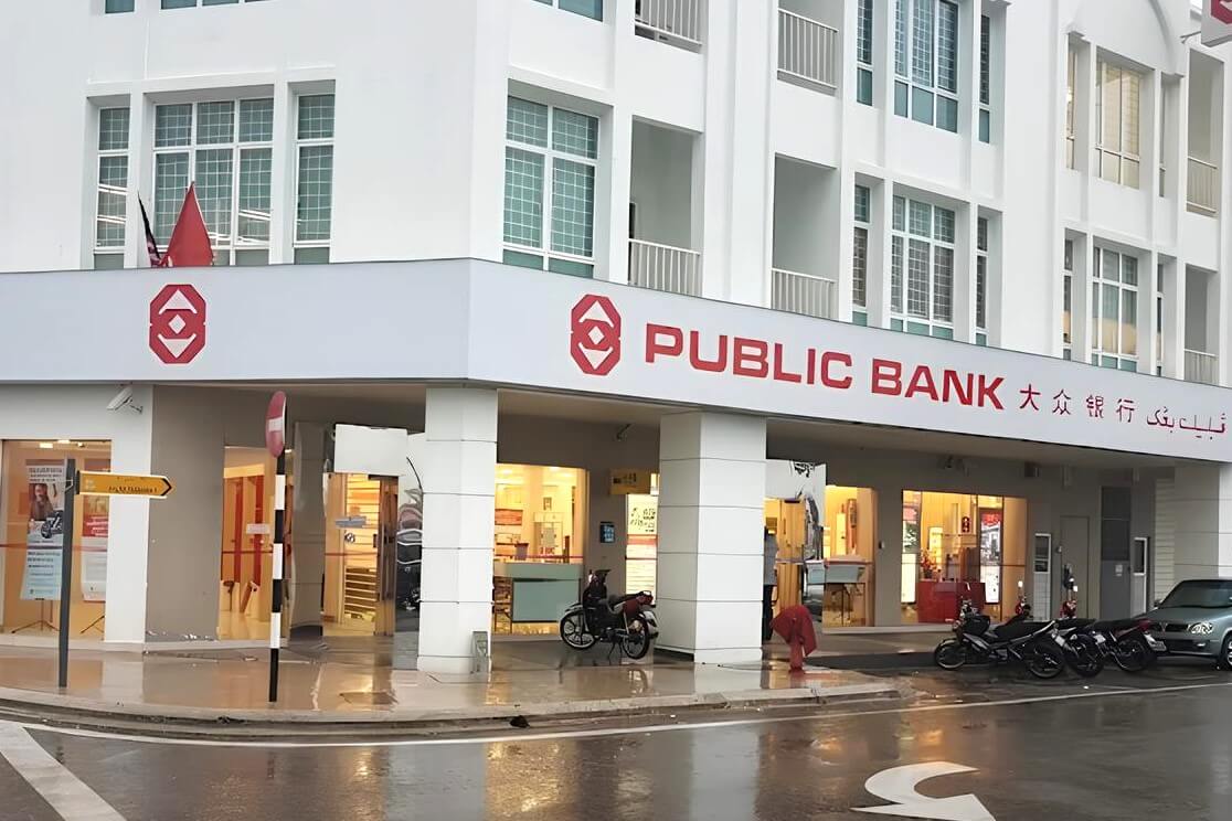 Bahagian luar bangunan Public Bank Tanah Merah pada hari hujan. Nama bank itu jelas dipaparkan dalam warna merah pada papan tanda putih di tingkat satu dalam tiga bahasa: Inggeris, Mandarin dan Arab. Bangunan ini ialah bangunan tiga tingkat berwarna putih dengan pintu masuk bertiang