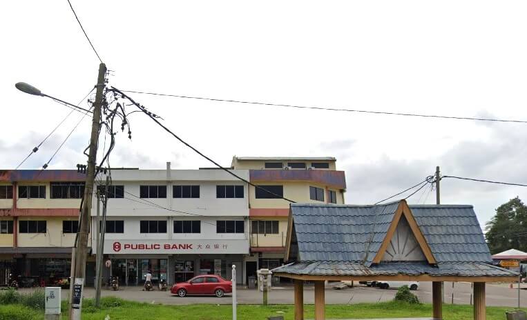 Gambar luaran menunjukkan cawangan Public Bank di Chaah, Malaysia, di bawah langit yang cerah dan mendung. Tumpuan utama ialah bangunan putih dan kuning tiga tingkat yang menempatkan bank di tingkat bawah. Logo merah bank dan nama "PUBLIC BANK" jelas kelihatan pada papan tanda di atas pintu masuk, dengan aksara Cina di sebelah kanan. Sebuah kereta sedan merah kecil diletakkan di jalan di hadapan bangunan itu