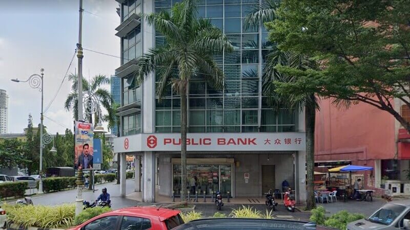 Pemandangan hadapan cawangan Public Bank di Jalan Tun Sambanthan pada waktu siang. Bangunan bertingkat ini mempunyai fasad kaca dengan papan tanda merah dan putih. Di hadapan bangunan terdapat pokok kelapa sawit/pohon hiasan dengan kenderaan yang lalu-lalang.