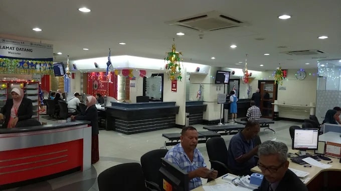 Bahagian dalam bangunan pejabat moden KWSP Alor Setar. Di latar depan, orang ramai, kebanyakannya lelaki, duduk di meja, nampaknya bekerja atau dilayan oleh kakitangan pejabat. Pejabatnya kemas, dengan meja dan kerusi yang tersusun rapi. Papan tanda di latar belakang berbunyi "Selamat Datang" dalam bahasa Melayu dan Inggeris