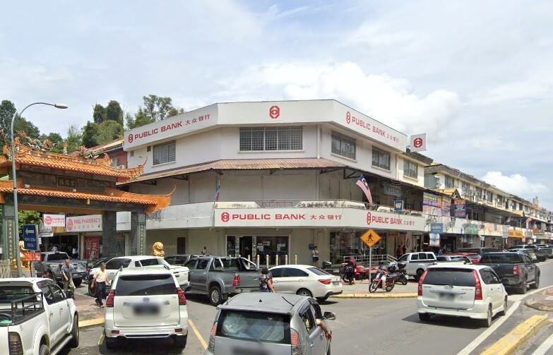 Gambar luar Public Bank Donggongon, sebuah bangunan putih tiga tingkat dengan papan tanda merah di tengah-tengah jalan komersial. Jalan ini sibuk dengan kereta yang parkir dan bergerak, dan pintu pagar tradisional Cina kelihatan di sebelah kiri