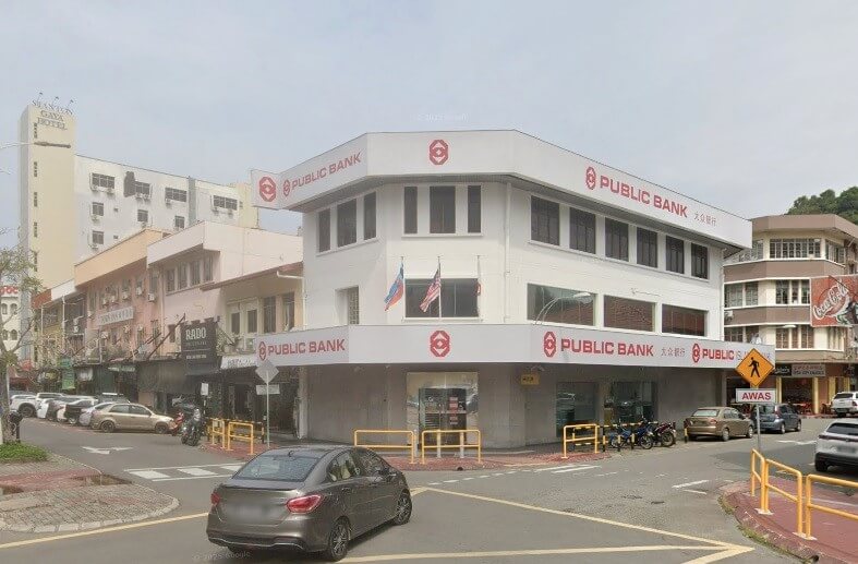 Bangunan Public Bank Jalan Pantai tiga tingkat berwarna putih dengan papan tanda merah di sudut jalan. Terdapat kereta yang diletakkan dan memandu berdekatan, dan papan tanda lintasan pejalan kaki kelihatan di sebelah kanan