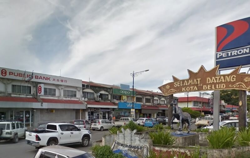 Pemandangan jalan siang hari di Kota Belud, Malaysia, menunjukkan deretan rumah kedai dengan papan tanda Public Bank dan stesen minyak Petron. Di latar depan, terdapat papan tanda selamat datang yang berbunyi "SELAMAT DATANG KE KOTA BELUD" bersebelahan dengan patung kuda hitam