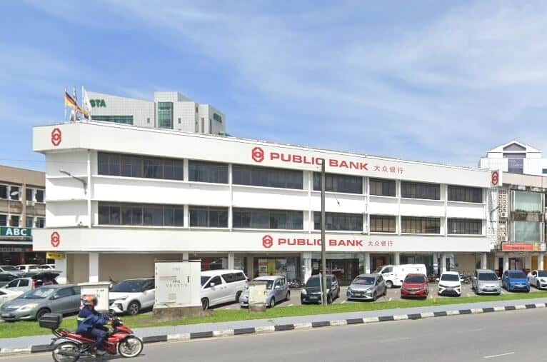 Pemandangan jalan luar bangunan Public Bank Berhad berwarna putih di Jalan Padungan, dengan kawasan parkir di hadapan dan kelihatan kereta serta motosikal