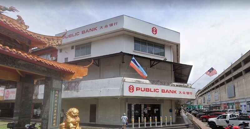 Bangunan Public Bank Donggongon dengan papan tanda “Public Bank 大众银行”, dihiasi bendera Malaysia dan bendera Sabah, terletak di kawasan perniagaan dengan sebuah bangunan berunsur seni bina Cina di sebelah kiri.