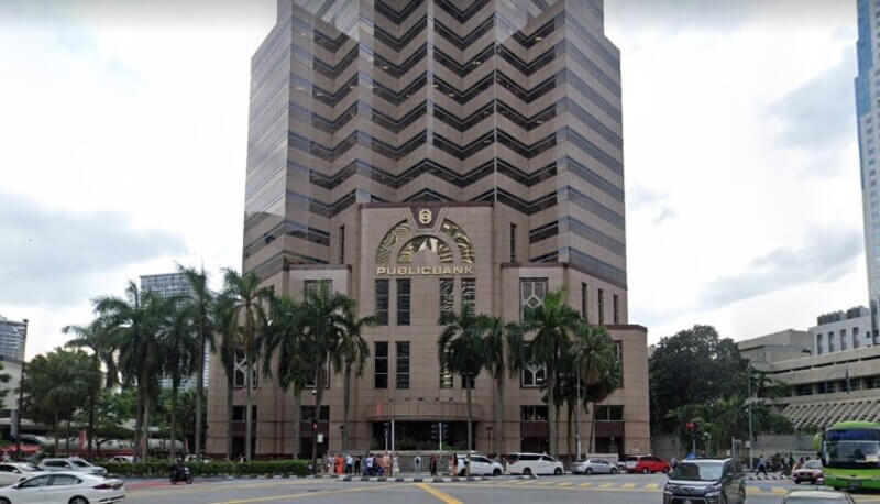 Pandangan hadapan Bangunan Menara Public Bank di Kuala Lumpur City Main Office dengan pokok kelapa sawit di bahagian hadapan dan jalan raya yang sibuk.