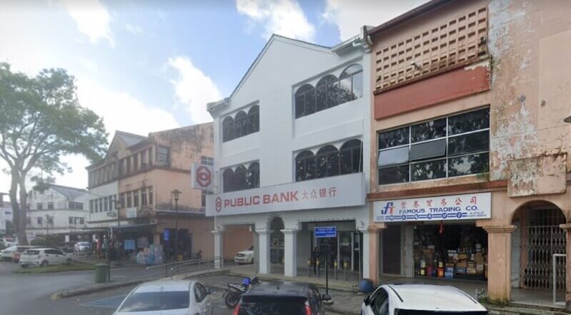 Bangunan cawangan Public Bank Mukah berwarna putih di deretan rumah kedai Mukah dengan papan tanda berhuruf merah dan tulisan Cina.