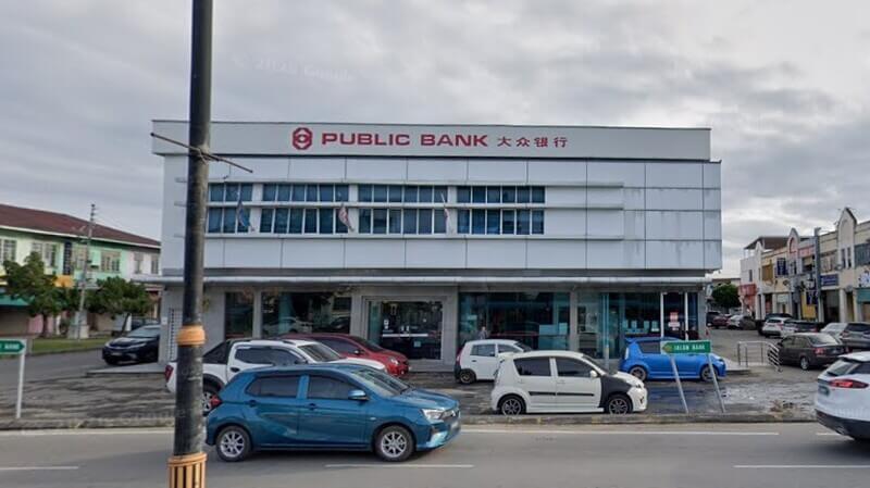 Pemandangan dari luar bangunan cawangan Public Bank Papar dua tingkat berpanel putih dengan papan tanda "PUBLIC BANK" dan tulisan Cina. Beberapa kenderaan diletakkan di hadapan bangunan di sepanjang jalan raya bawah cuaca agak berawan.