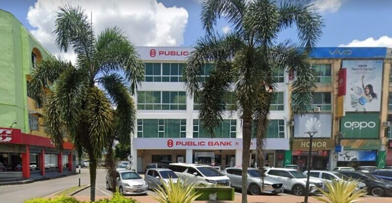 Bangunan cawangan Public Bank Sarikei empat tingkat bertembok putih dengan papan tanda merah, dikelilingi pokok kelapa sawit hiasan dan tempat letak kereta.