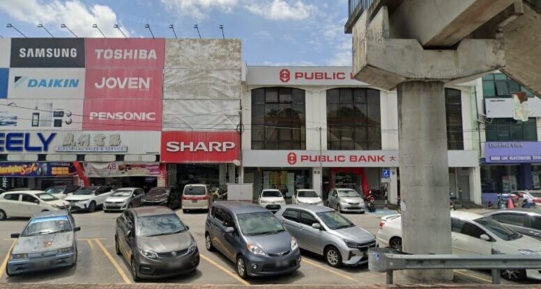 Pemandangan hadapan cawangan Public Bank Simpang Ampat dan kedai barangan elektrik di tepi jalan barisan kedai, dengan kereta diletakkan di petak letak kereta dan tiang struktur konkrit di latar hadapan.