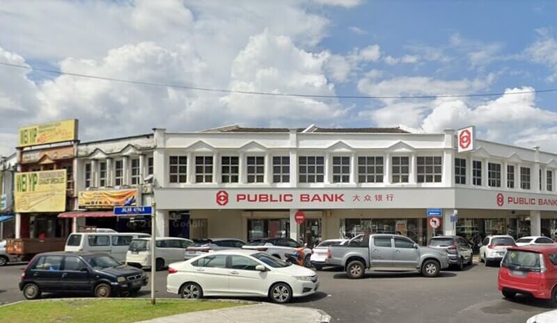 Pemandangan luar cawangan Public Bank Taman Cheras dua tingkat berwarna putih di tepi jalan dengan papan tanda kedai jiran, deretan kenderaan yang diparkir di hadapan, dan langit berawan.