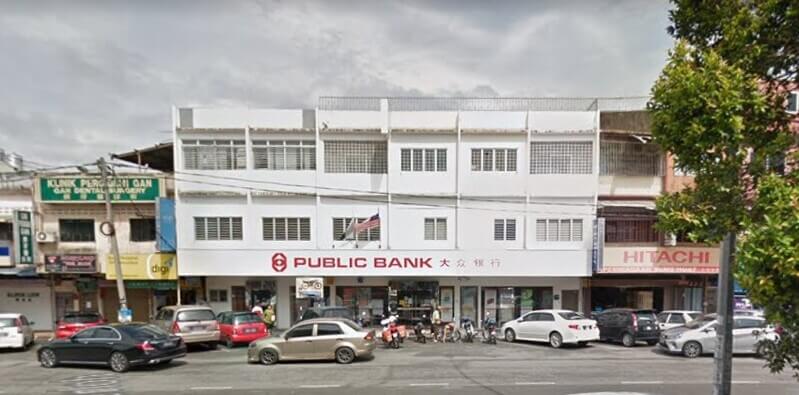 Pandangan hadapan bangunan cawangan Public Bank Triang di barisan kedai Triang dengan tempat letak kereta di hadapannya.