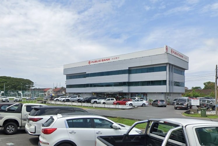 Bangunan tiga tingkat Public Bank dengan bahagian luar kelabu dan putih moden, dikelilingi oleh tempat letak kereta yang besar penuh dengan kereta dan SUV. Bangunan itu mempunyai logo dan nama bank berwarna merah di bahagian atas muka bangunan
