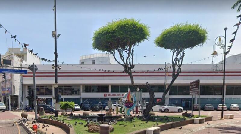 Pemandangan luar bangunan Public Bank Melaka di tepi jalan dengan kawasan lanskap hiasan, pokok-pokok bercabang tinggi, dan kawanan burung merpati di sekelilingnya.