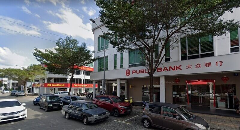 Pandangan luaran bangunan cawangan Public Bank Taman Melaka Raya dan AmBank di tepi jalan kawasan perniagaan dengan deretan kenderaan yang diletakkan di hadapannya.