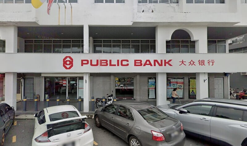 Pandangan hadapan cawangan Public Bank di Bandar Baru Air Itam yang memaparkan papan tanda bank berwarna merah dan putih, laluan pejalan kaki di hadapan kedai, serta beberapa buah kereta dan motosikal yang diletakkan di kawasan parkir.