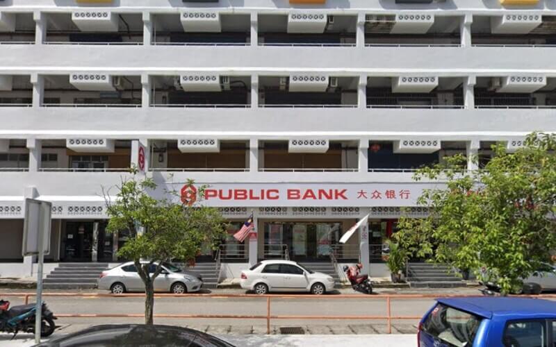 Pemandangan luar cawangan Public Bank di Lebuh Macallum di tingkat bawah sebuah bangunan bertingkat, dengan beberapa buah kereta diletakkan di hadapan papan tanda bank.