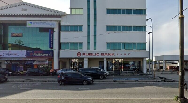 Pandangan hadapan bangunan cawangan Public Bank Port Klang bertingkat di tepi jalan raya dengan papan tanda berhuruf merah dan barisan kenderaan diletakkan di hadapannya.