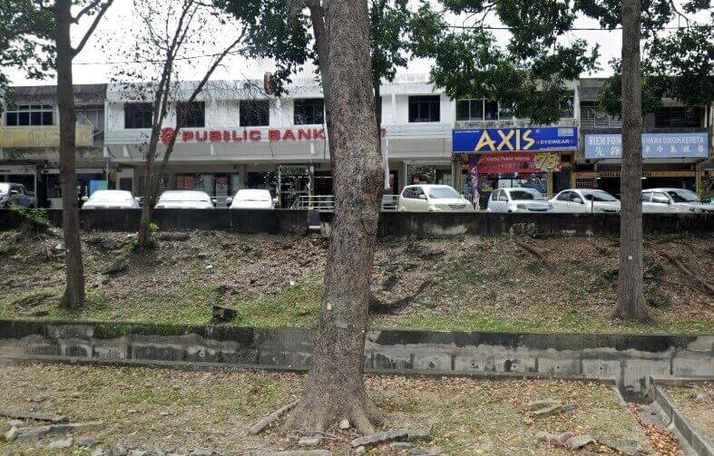 Foto luar yang diambil dari seberang parit atau tembok penahan rendah menunjukkan deretan rumah kedai dua tingkat. Tumpuan utama adalah bangunan di tengah, yang menempatkan cawangan Public Bank dengan logo merah dan putih serta nama boleh dilihat di tingkat satu. Di sebelah kanan bank adalah sebuah kedai dengan papan tanda besar berwarna kuning dan biru untuk Axis Eyewear. Kedai-kedai lain yang lebih kecil kelihatan lebih jauh ke kiri dan kanan