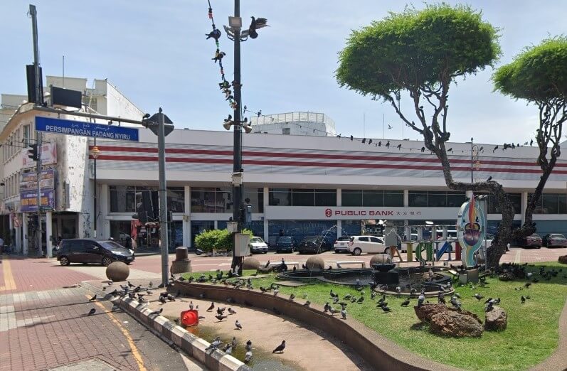 Pemandangan jalan cawangan Public Bank di Melaka, Malaysia, terletak di seberang dataran sibuk dan pulau trafik kecil. Bangunan bank itu besar, putih, dan mempunyai jalur merah yang menyerlah dengan namanya di tengah. Di latar depan, papan tanda lalu lintas menghala ke "Persimpangan Padang Nyiru."