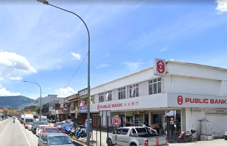 Cawangan Public Bank di kawasan komersial yang sibuk di Nilai, Malaysia. Bangunan dua tingkat itu kebanyakannya berwarna putih dengan kemasan merah dan memaparkan logo bank (bentuk "P" merah yang digayakan dengan jelas). Papan tanda utama bertulis "PUBLIC BANK" dalam huruf merah besar, dengan aksara Cina di bawah, mengenal pasti ia sebagai Public Bank Berhad di Malaysia