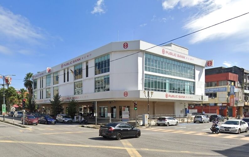 Bangunan putih tiga tingkat pada hari yang cerah di persimpangan yang sibuk. Ia adalah cawangan Public Bank, dengan logonya (bulatan merah dengan 'P' bergaya di dalam) dan nama dalam skrip Melayu dan Mandarin boleh dilihat di bahagian luar