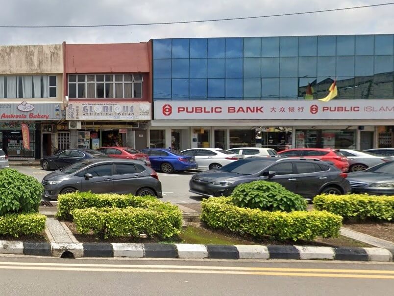 Foto deretan rumah kedai dari aras jalan. Bangunan utama di sebelah kanan adalah cawangan Public Bank, yang mempunyai fasad yang besar dan moden dengan kaca pantulan biru di tingkat atas dan papan tanda putih bertulis "PUBLIC BANK" dan "PUBLIC ISLAMIC BANK" dalam bahasa Inggeris dan Cina untuk nama bank. Di sebelah kiri bank itu terdapat dua perniagaan lain: "GLOROUS ATTIRIES TAILORING" dan "Ambrosia Bakery."