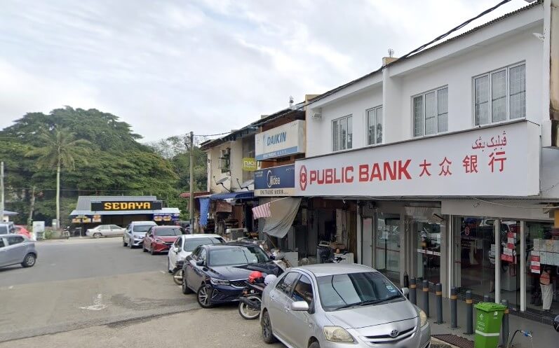 Pemandangan jalan menunjukkan bahagian luar cawangan Public Bank di Benta, Malaysia. Papan tanda merah putih bank itu jelas kelihatan di sebuah rumah kedai dua tingkat. Beberapa kereta diletakkan selari dengan kaki lima di hadapan bank dan pelbagai perniagaan di sekitarnya, termasuk sebuah kedai Daikin dan sebuah lagi kedai Midea