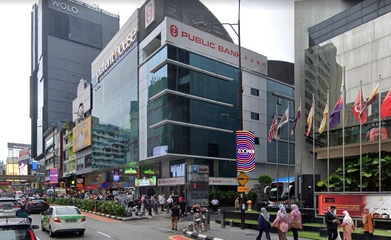 Bangunan kaca dan konkrit bertingkat di sudut jalan bandar yang sibuk, dengan logo 'PUBLIC BANK' merah dan putih yang menonjol di bahagian atas. Bangunan ini merupakan sebahagian daripada persekitaran bandar yang padat dengan bangunan bertingkat tinggi yang lebih gelap berlabel 'WOLO' boleh dilihat di sebelah kiri