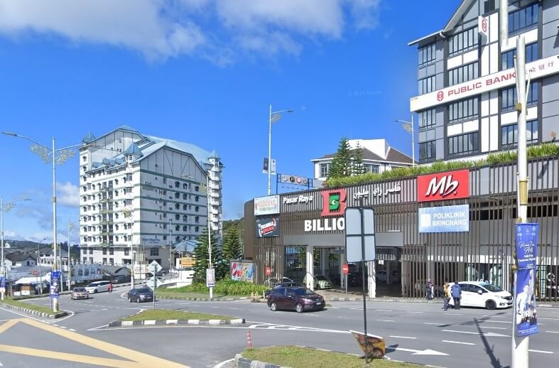 Pemandangan jalan siang hari di Brinchang, Malaysia, menunjukkan kawasan komersial moden. Di sebelah kanan, bangunan bertingkat memaparkan papan tanda "PUBLIC BANK" di tingkat atas dan pasar raya "BILLION", "MB" (Marrybrown) dan "POLIKLINIK BRINCHANG" di tingkat bawah