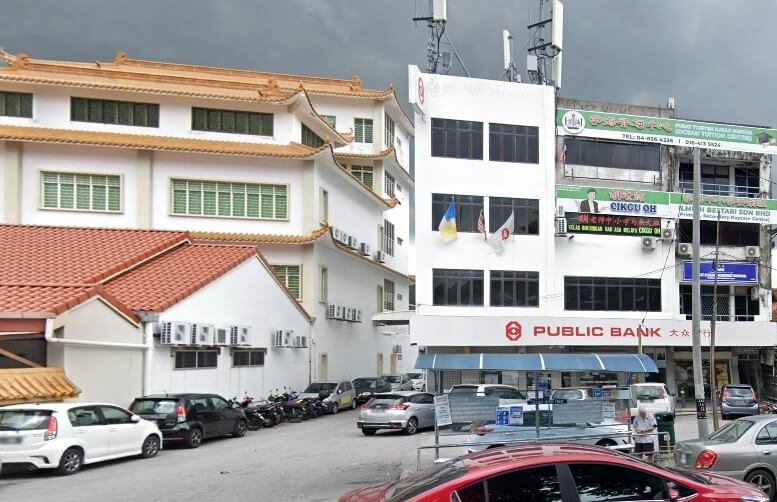 Pemandangan jalan yang menunjukkan bangunan cawangan Public Bank di Jalan Air Itam di Malaysia. Bangunan bank bertingkat berwarna putih ini terletak di sebelah kanan, bersebelahan dengan kuil atau institusi putih gaya tradisional yang besar dengan bumbung coklat di sebelah kiri