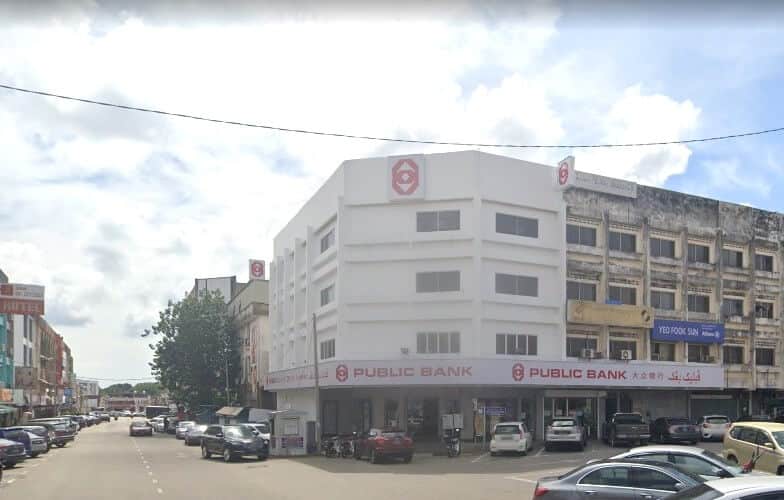 Foto Pejabat Public Bank Mentakab dari aras jalan, bangunan sudut putih empat tingkat dengan logo Bank Komersial dan nama boleh dilihat pada fasadnya. Bangunan ini terletak di jalan yang sibuk dengan kereta diletakkan di hadapan dan di sebelahnya, dan bangunan lama gaya rumah kedai bertingkat bersebelahan antara satu sama lain di bawah langit yang mendung