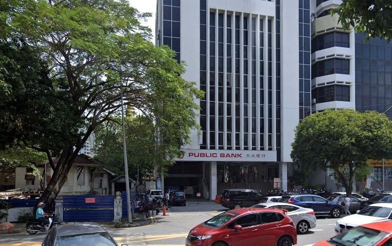 Foto Pejabat Public Bank Pulau Tikus dari aras jalan, bangunan putih tinggi dengan tingkap menegak. Nama bank ditulis dengan huruf merah di atas pintu masuk. Jalan yang sibuk dengan beberapa kereta dan beberapa pembinaan di sebelah kiri kelihatan di latar depan