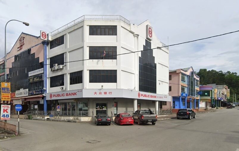 Bangunan komersial empat tingkat putih dan biru di sudut jalan ditandai dengan papan tanda besar sebagai Pejabat PUBLIC BANK Raub, dengan huruf Cina "大众银行" pada muka depannya. Beberapa buah kereta diletakkan di hadapan bangunan di atas jalan berturap. Bank ini diapit oleh rumah kedai kecil berwarna-warni di kedua-dua belah pihak