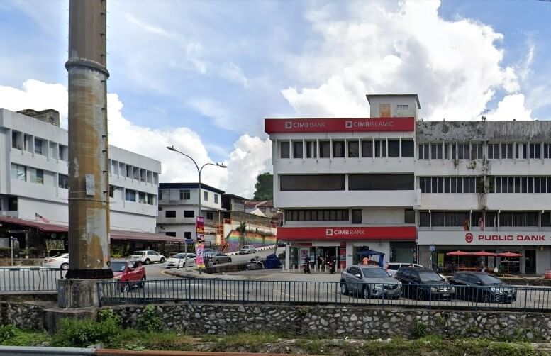 Pemandangan jalan beberapa bangunan komersial di Taman Connaught. Di latar depan, tiang atau cerobong konkrit yang besar dan lapuk berdiri dengan bangga di sebelah kiri. Dinding batu rendah memisahkan latar depan dari jalan, dan parit atau aliran kecil yang ditumbuhi kelihatan di bahagian bawah. Bangunan mendominasi pandangan ke kanan dan tengah-kanan. Paling menonjol ialah bangunan putih empat tingkat dengan papan tanda merah besar yang mewakili cawangan CIMB Bank dan Public Bank