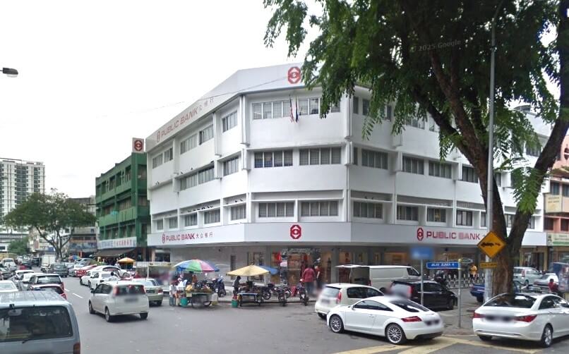 Pemandangan jalan cawangan Public Bank di Taman Maluri. Tumpuan utama ialah bangunan putih empat tingkat di sudut jalan yang sibuk. PUBLIC BANK dipamerkan dengan jelas dalam huruf merah pada fasad, termasuk logo bulatan merah dengan huruf "P" dan "B" di sudut atas dan di atas pintu masuk di tingkat bawah