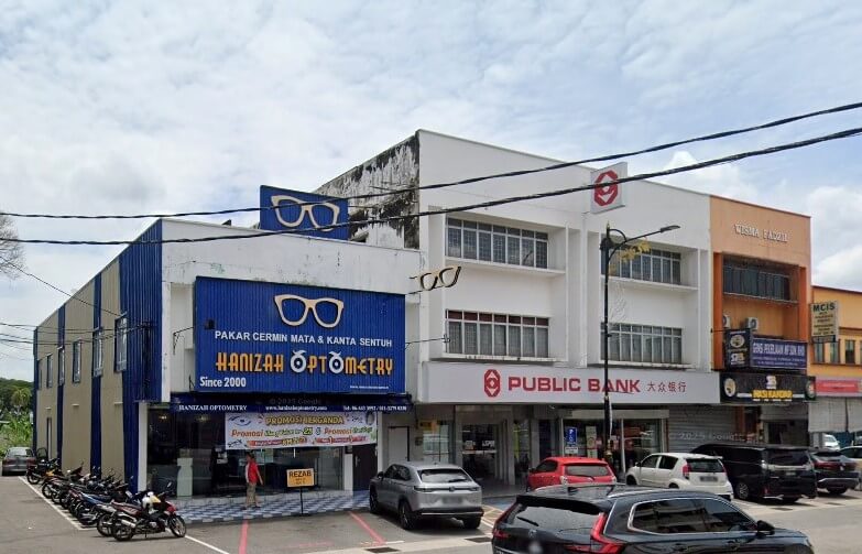 Pemandangan jalan deretan bangunan komersial. Di sebelah kiri, bangunan biru-putih menempatkan Hanizah Optometri, ditandai dengan papan tanda biru besar dengan huruf putih bertulis "PAKAR CERMIN MATA & KANTA SENTUH". Di sebelah kanan, sebuah bangunan berwarna putih memaparkan cawangan Public Bank di tingkat bawah, dengan logo merah-putih dan huruf merahnya dipaparkan dengan jelas. Tingkap di tingkat atas kelihatan di atas tebing