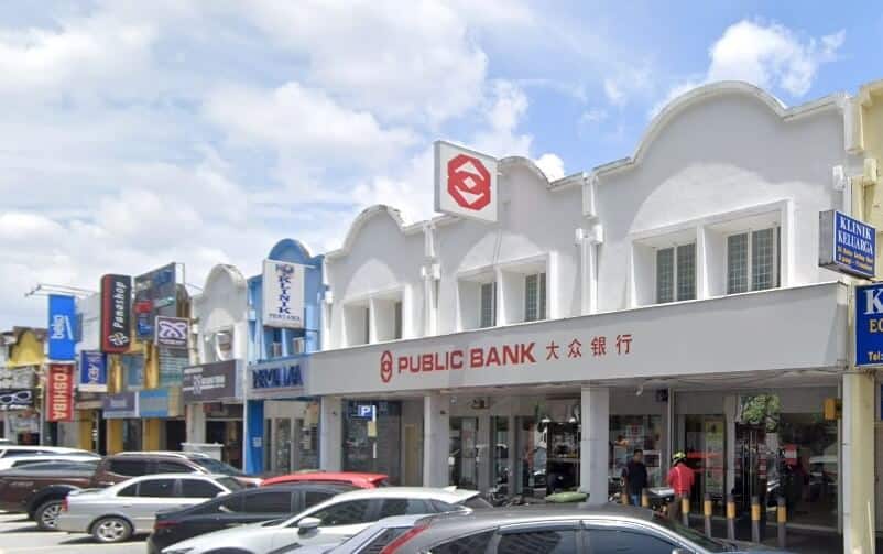 Pemandangan jalan siang hari yang cerah di cawangan Public Bank di Wangsa Maju, Kuala Lumpur, Malaysia. Bank itu terletak di tingkat bawah sebuah rumah kedai komersial dua tingkat berwarna putih dengan fasad melengkung dan papan tanda merah putih yang memaparkan logo dan nama bank dalam aksara Inggeris dan Mandarin