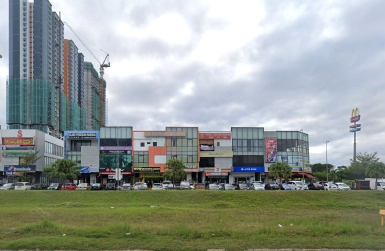 Gambar jalanan deretan rumah kedai komersil moden pelbagai warna di Kota Warisan, Malaysia, menampilkan perniagaan seperti Affin Bank dan 7-Eleven, dengan bangunan pangsapuri bertingkat tinggi yang sebahagiannya dibina kelihatan di belakangnya di sebelah kiri, dan papan tanda McDonald's di hujung kanan