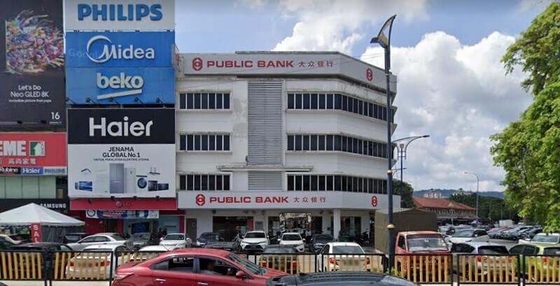 Pandangan hadapan bangunan cawangan Public Bank Ampang empat tingkat dengan papan tanda kedai barangan elektrik di sebelahnya dan kawasan tempat letak kereta yang dipenuhi kenderaan.