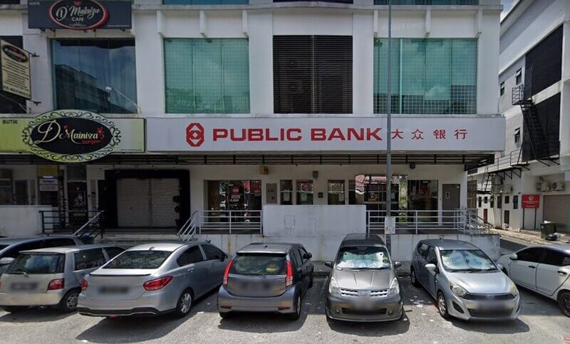 Pandangan hadapan cawangan Public Bank Bandar Kinrara dengan papan tanda merah dan putih, kedai D'Mainiza di sebelah kiri, serta deretan kereta yang diletakkan di petak letak kereta di hadapannya.