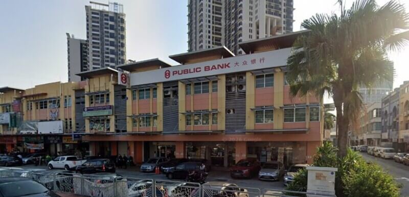 Pandangan luar bangunan cawangan Public Bank Kelana Jaya di kawasan rumah kedai dengan papan tanda perbankan, kawasan tempat letak kereta, dan bangunan kediaman tinggi di latar belakang.
