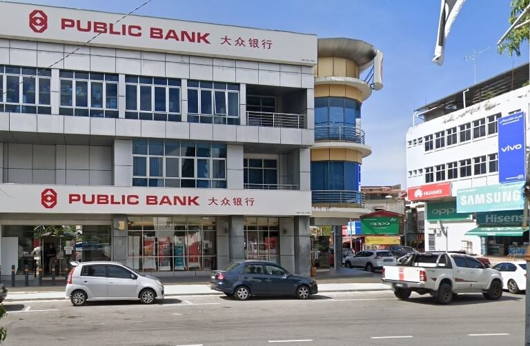 Bangunan moden bertingkat dengan perkataan "PUBLIC BANK" dalam huruf merah besar pada muka depan kelabu muda. Beberapa kereta parkir di hadapan bangunan itu, dan pelbagai bangunan komersial dengan papan iklan kelihatan di sebelah kanan