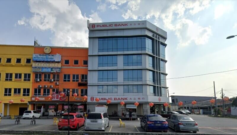 Pandangan hadapan bangunan cawangan Public Bank di Rawang dengan tempat letak kereta dan kedai-kedai bersebelahan di bawah langit cerah.