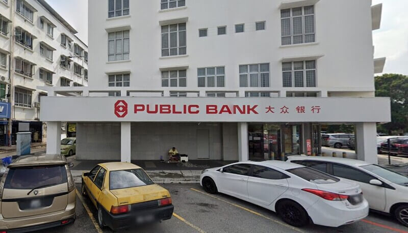 Pandangan hadapan bangunan cawangan Public Bank Shah Alam dengan papan tanda merah dan beberapa kenderaan di petak meletak kereta.