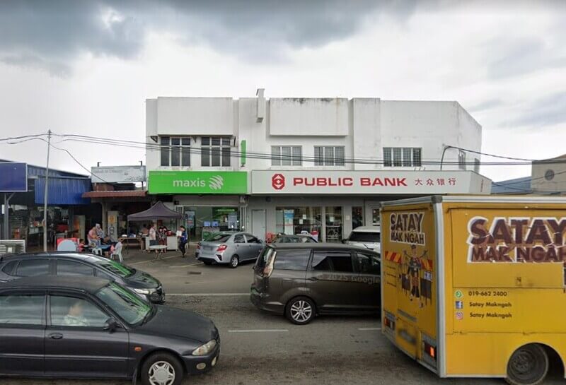 Pemandangan dari luar bangunan dua tingkat yang menempatkan cawangan Public Bank Sungai Pelek dan pusat perkhidmatan Maxis di tepi jalan utama dengan beberapa kenderaan diletakkan di hadapannya.